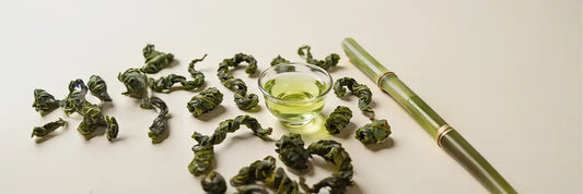 A close-up of tamaryokucha loose leaf green tea showing its distinctive small comma-shaped curled leaves in a natural light, with a pale yellow-green brewed cup alongside.