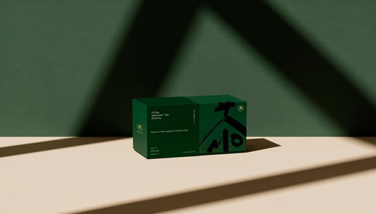A dark green rectangular box of Tea sits on a beige surface against a dark green wall, with dramatic diagonal shadows cast across the scene.