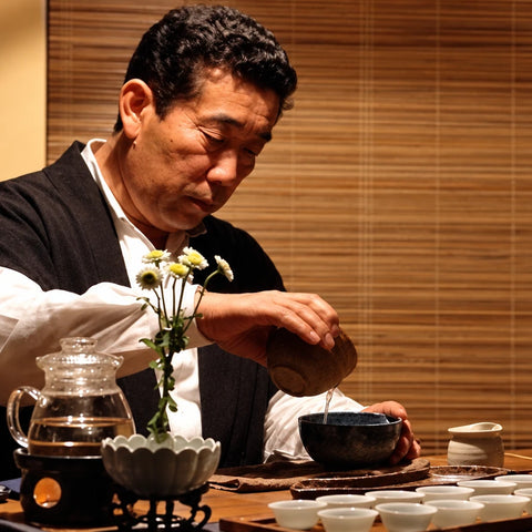 Chanoka Farmer in traditional Japanese attire pouring tea from a glass teapot into small white cups on a wooden table, with a small vase of white flowers in the foreground.