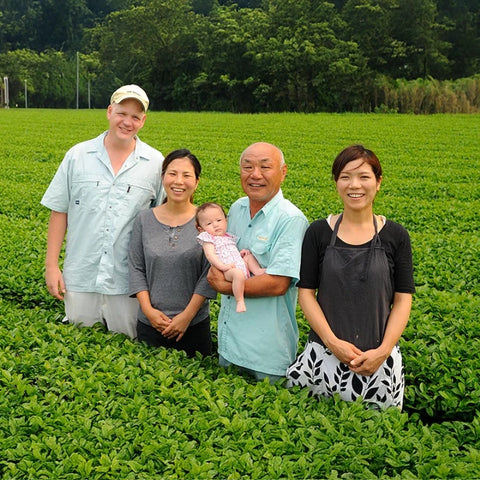 Chiran farmer family standing together in a lush green tea field, with one person holding a baby. The group is dressed in light-colored casual and work clothing, set against a background of dense trees.