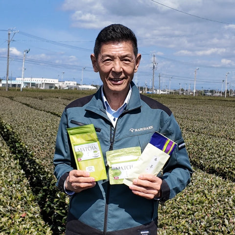 Masuda farmer wearing a teal and black Mardaki jacket stands in a tea plantation holding three packages of M-Tea Matcha green tea.
