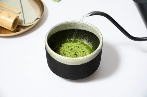 A ceramic matcha tea bowl filled with green matcha powder, with hot water being poured into it from a black gokuroh kettle on a white surface.
