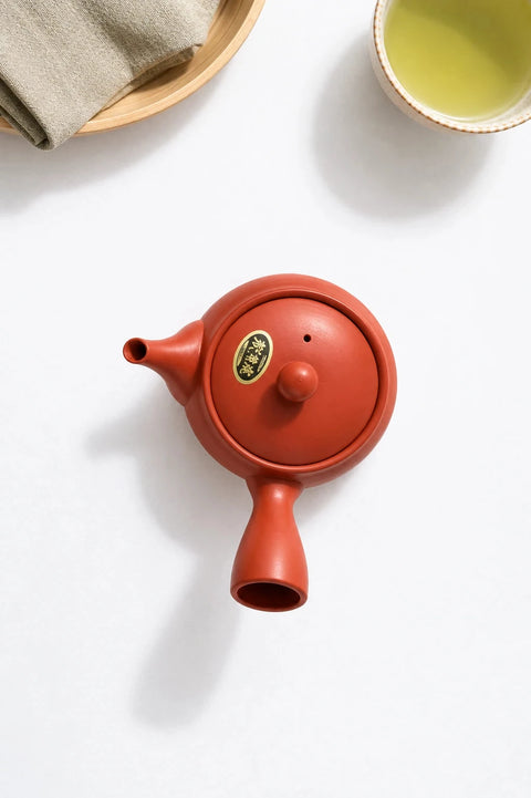A red ceramic teapot with a matching lid and a small round gold logo badge, photographed on a light beige surface with tea cups partially visible around it.