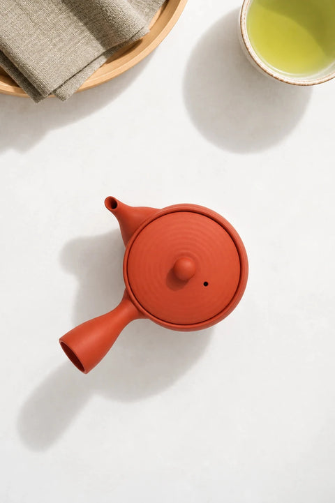 A terracotta red clay teapot with a matching lid, styled on a white surface with a cup of green tea, a folded beige cloth, and a wooden tray partially visible.